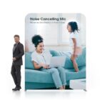 A woman sits on a teal sofa with a laptop, wearing earphones. A child joyfully jumps beside her. A man in a suit walks past, and text above them reads: Noise Cancelling Mic - Never be disturbed by ambient noise.