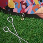 Close-up image of green artificial grass with two metal pegs on it. Above, there is a colorful illustration of people in action, wearing sports attire. A black border with an attached ring separates the grass and the illustration.