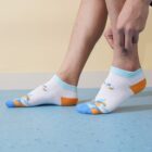 A person wearing colorful, mismatched socks adjusts one of them. The socks are white with blue, orange, and gray patterns, featuring small shapes. The person is standing on a blue speckled floor against a light-colored wall.