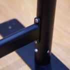 Close-up of a black metal bracket connecting a horizontal bar to a vertical pole. The bracket is secured with two screws, and the structure is mounted on a wooden surface.