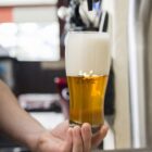 A hand holds a glass of beer with a thick, foamy head at a bar or brewery.