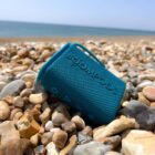 A teal portable speaker on a rocky beach with the sea in the background.