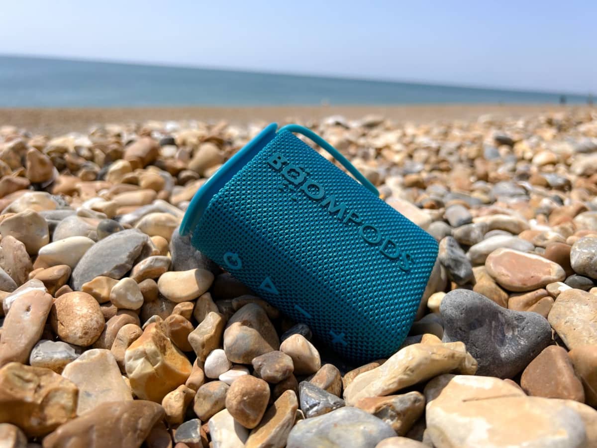 A teal portable speaker on a rocky beach with the sea in the background.