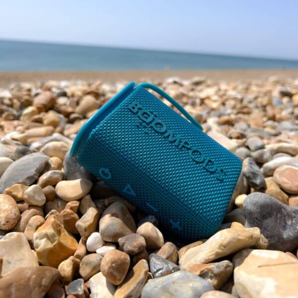 Blue portable speaker on rocky beach with sea and sky in the background.