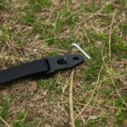 A black strap is secured to the ground with a metal stake on patchy grass. The stake is inserted through a hole in the straps plastic end. The scene suggests outdoor anchoring or securing equipment.