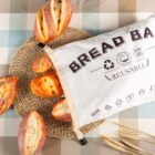 Several crusty bread loaves are partially in a white reusable bread bag with text and symbols, on a woven mat. The background is a checkered, neutral-toned tablecloth, with three wheat stalks beside the bag.