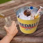 A person is pouring beer from a bottle into a glass. Three bottles of beer are chilled in ice inside a metal bucket labeled Medalla Light on a wooden table.