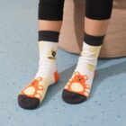 Child sitting with legs crossed, wearing white socks featuring orange bears and black toe sections. The socks have a brand logo and text. Theyre seated on a beige surface with a light blue, speckled floor.