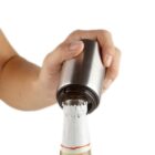 A hand uses a metal bottle opener to remove the cap from a sealed bottle, which is wrapped in white foil. The background is plain white.