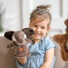 A young girl wearing a tiara smiles while holding a plush elephant puppet. She is seated on a sofa, wearing a light blue dress. Another plush puppet is partially visible on the right. The background is softly blurred.