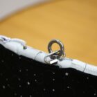 A close-up of a fabric with a black background featuring moon and star patterns. The fabric is attached to a curved metal frame with a loop and latch mechanism, set against a blurred wooden surface.