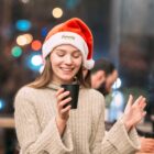 Woman in a Santa hat and a beige sweater smiles while holding a black cup. The background is softly blurred with colorful lights, suggesting a festive atmosphere.