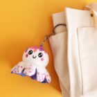 A cute plush jellyfish keychain with a smiling face, pink and purple patterns, is attached to the handle of a beige bag. The background is bright yellow.