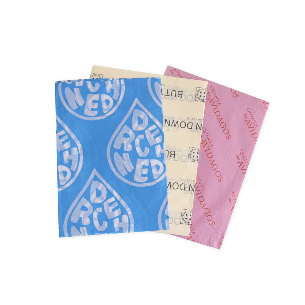 Three sheets of tissue paper are displayed. One is blue with a bold DENCHED pattern in teardrop shapes, another is off-white with cartoon faces, and the third is pink with AMIGOS printed in red.