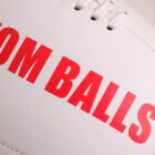 Close-up of a white ball with red text partially visible, showing the words OM BALLS. Stitching is visible at the top. The image focuses on the texture and detail of the balls surface and lettering.