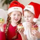 Two children wearing red Santa hats, with Embroidery Effect written on them, are joyfully looking at decorated cupcakes in their hands. They are sitting together indoors, sharing a festive moment.