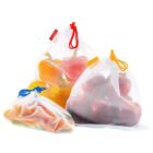 Three mesh produce bags filled with fresh vegetables and fruit: one with green and orange carrots, one with orange and yellow bell peppers, and one with red apples. Each bag has a drawstring in blue, red, or yellow.