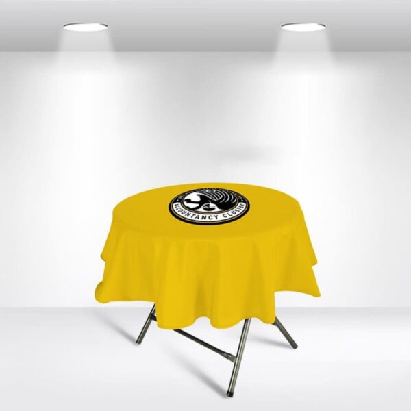 A yellow table cover with a black and white logo in the center is draped over a round table. The table sits under two spotlights in a minimalist room with white walls and floor.