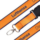 Close-up of an orange lanyard with the word Lufthansa printed in black. It features a black plastic buckle and a silver metal clasp for attaching items.