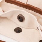Close-up of a beige fabric bag featuring a pair of metal snap buttons. The top button is open, highlighting the leather trim on the flap. The texture of the fabric and the stitching details are visible.