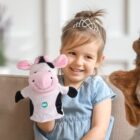 A young child wearing a tiara and a light blue dress is smiling while holding a cow hand puppet with a GO logo. The child is sitting on a sofa, and soft toys are around. The room has a light and cozy atmosphere.
