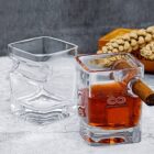 Two uniquely shaped square glasses are shown. One is empty, and the other contains a brown liquid with a cigar rested in a side notch. A wooden box with dried flowers is in the background on a gray surface.