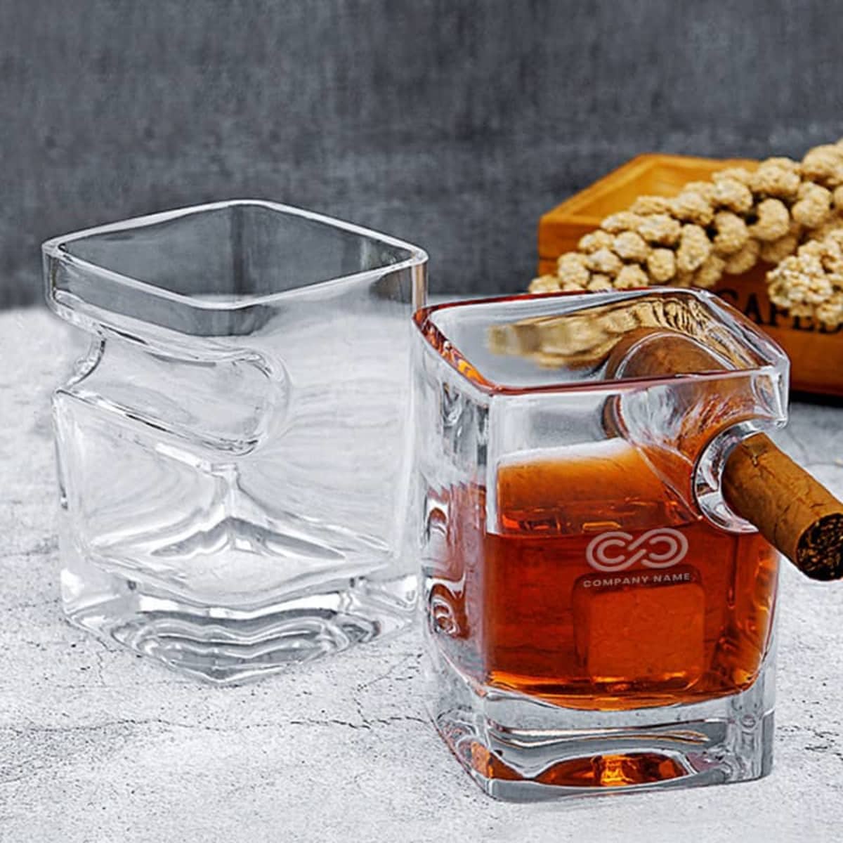 Two uniquely shaped square glasses are shown. One is empty, and the other contains a brown liquid with a cigar rested in a side notch. A wooden box with dried flowers is in the background on a gray surface.