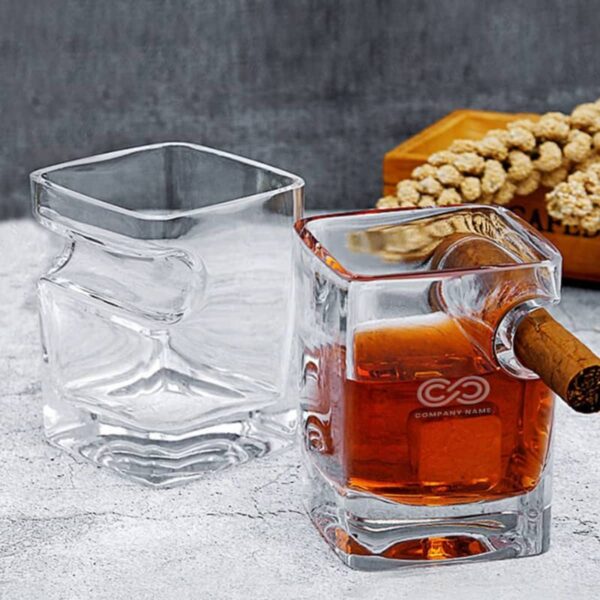 Two square-shaped glass tumblers are shown. One is empty, and the other contains brown liquid and a cigar resting on its rim. A wooden tray with dried plants is in the background on a gray surface.
