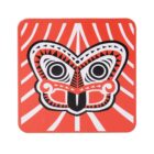 A square, red coaster features a stylized, symmetrical mask design in black and white. The background has white, radiating lines, creating a dynamic contrast with the bold central image.