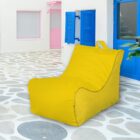 A modern outdoor scene with a bright yellow bean bag chair on a stone-patterned patio. The background features a white building with blue doors and shutters, and a pink-trimmed window, creating a vibrant, Mediterranean vibe.
