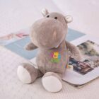 A soft, gray plush hippopotamus toy sits on a bed with a white, textured blanket. The hippo has embroidered eyes, small white ears, and Kids with a colorful handprint on its belly. An open magazine is in the background.