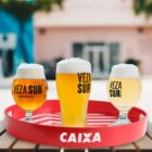 Three beers in different glasses sit on a red tray. Each glass has Veza Sur printed on it, showing varying beer colors from golden to amber. The background features a blurred outdoor setting with pastel buildings.