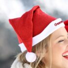 A person wearing a red and white Santa hat is smiling. The hat has a logo with red text. The background is blurry, giving a wintery feel.