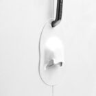 A minimalist coat hook designed like a melting white blob is mounted on a smooth, light-colored wall. A single black coat hanger is hooked onto it, creating a contrast with the white surface.