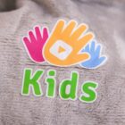Logo featuring three intertwined hands in pink, orange, and blue, with a play button in the center of the orange hand. The word Kids is written below in bright green letters on a gray textured background.