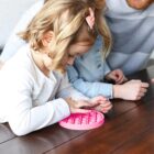 A young child with a pink hair bow plays with a pink pop-it toy on a wooden table. An adult and another child sit nearby, watching and engaging.