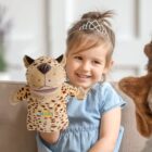 A smiling young girl with a tiara is sitting on a couch, holding a plush leopard puppet. She is wearing a blue dress and looking at another puppet to her right. The room is softly lit and decorated with neutral colors.