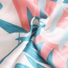 Close-up of a silky fabric with a colorful abstract leaf pattern featuring teal and pink shades. The material appears smooth and soft, with gentle folds and a slight sheen, creating a sense of movement and fluidity.