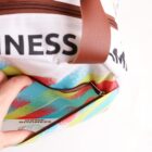 A colorful travel bag with zigzag patterns in blue, yellow, and red. The bag features brown handles and a partially open zipper compartment, with a glimpse of text on the side. A persons hand is holding the bag.