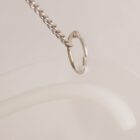 Close-up of a delicate silver necklace with a circular pendant, resting on a white, glossy surface. The pendant is attached to a fine chain, and the overall setting is minimalist, emphasizing the jewelrys simplicity and elegance.
