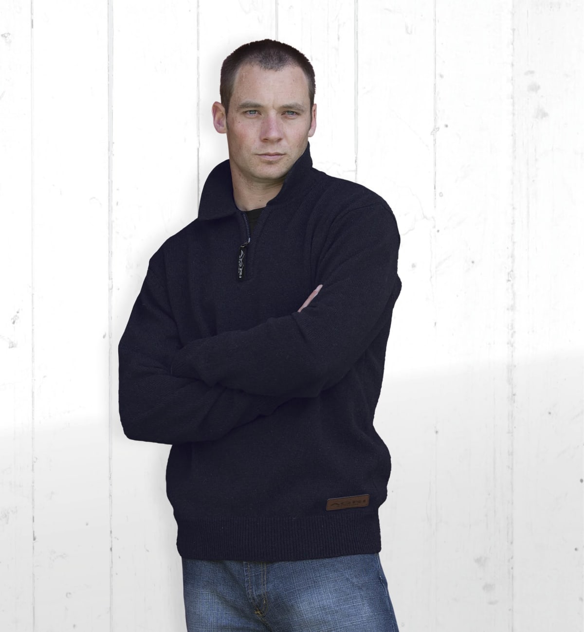 A person wearing a black sweater and blue jeans stands with arms crossed against a white wooden background, looking slightly to the side.