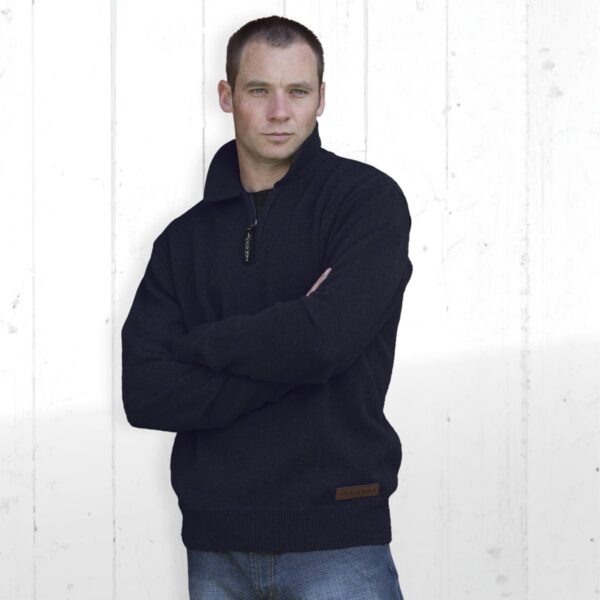 A person stands against a light-colored wooden wall, wearing a black zippered sweater and blue jeans. The person has short hair and is looking slightly to the side with a neutral expression, arms crossed.