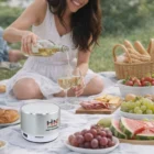Woman pours white wine at a picnic with food, fruit, and an Adler Bluetooth Speaker.