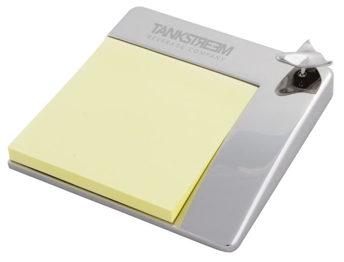 A silver metallic note holder with a pad of yellow sticky notes. It has a bottle opener on the top right corner and the text TANKSTREAM BEVERAGE COMPANY engraved on the top.