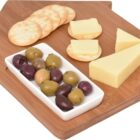 A wooden cheese board shaped like a house with a built-in spreader. It has various types of cheese, a small dish of assorted olives, and several round crackers arranged on it.