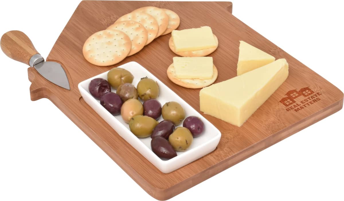 A wooden cheese board shaped like a house with a built-in spreader. It has various types of cheese, a small dish of assorted olives, and several round crackers arranged on it.
