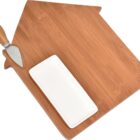 A wooden cutting board shaped like a house, featuring a small handle on one end. It has a white rectangular dish and a metal cheese knife with a wooden handle on it.