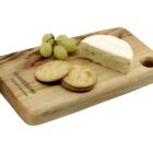 A wooden cheese board with a small bunch of green grapes, four round crackers, and a wedge of soft cheese. The board has a hole in one corner and engraved text.