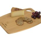 A bamboo cutting board with a wedge of cheese, a small bunch of grapes, and several round crackers neatly arranged. The board has an engraved logo in the corner and a handle for easy carrying.