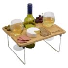 A wooden picnic table holds two glasses of white wine, a bottle, cheese, crackers, salami slices, and grapes. The table has foldable metal legs, and the Allianz logo is engraved on the surface.